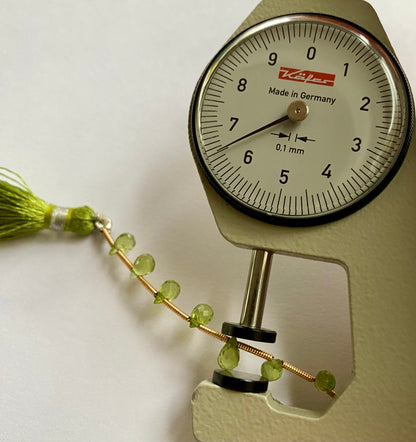 A close-up of a person measuring a peridot faceted briolette  3.7mm-to-4.4mm bead stone, with  dial gauge.