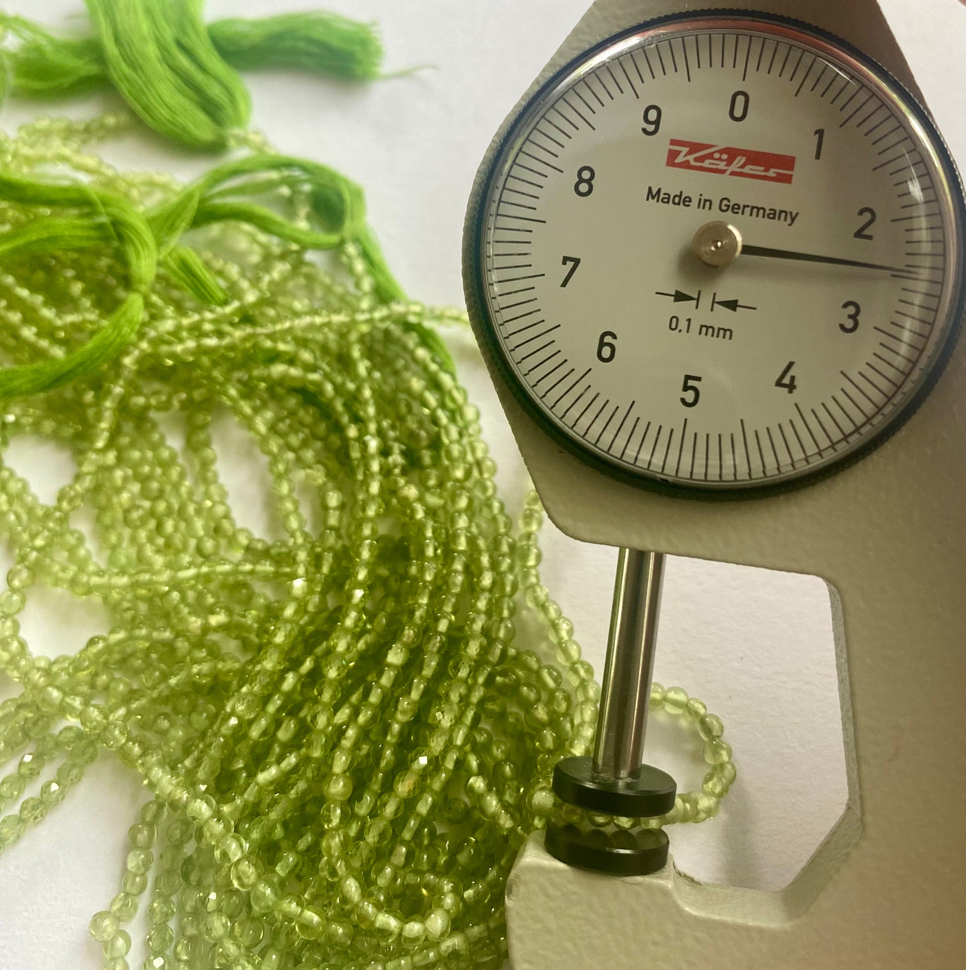 A person measures a green peridot smooth round stone, ranging from 2.5mm to 2.7mm, using a dial gauge measuring tool.
