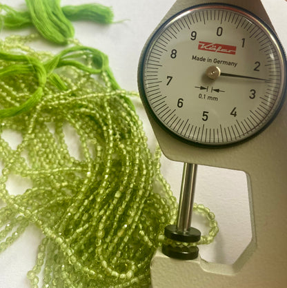 A person measures a green peridot smooth round stone, ranging from 2.5mm to 2.7mm, using a dial gauge measuring tool.
