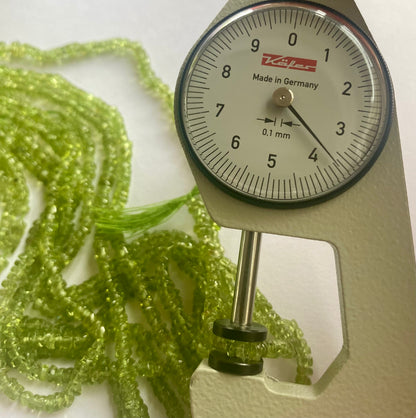 Close-up of a measuring dial gauge alongside smooth green peridot beads, ranging from 3.5mm to 4mm in size.