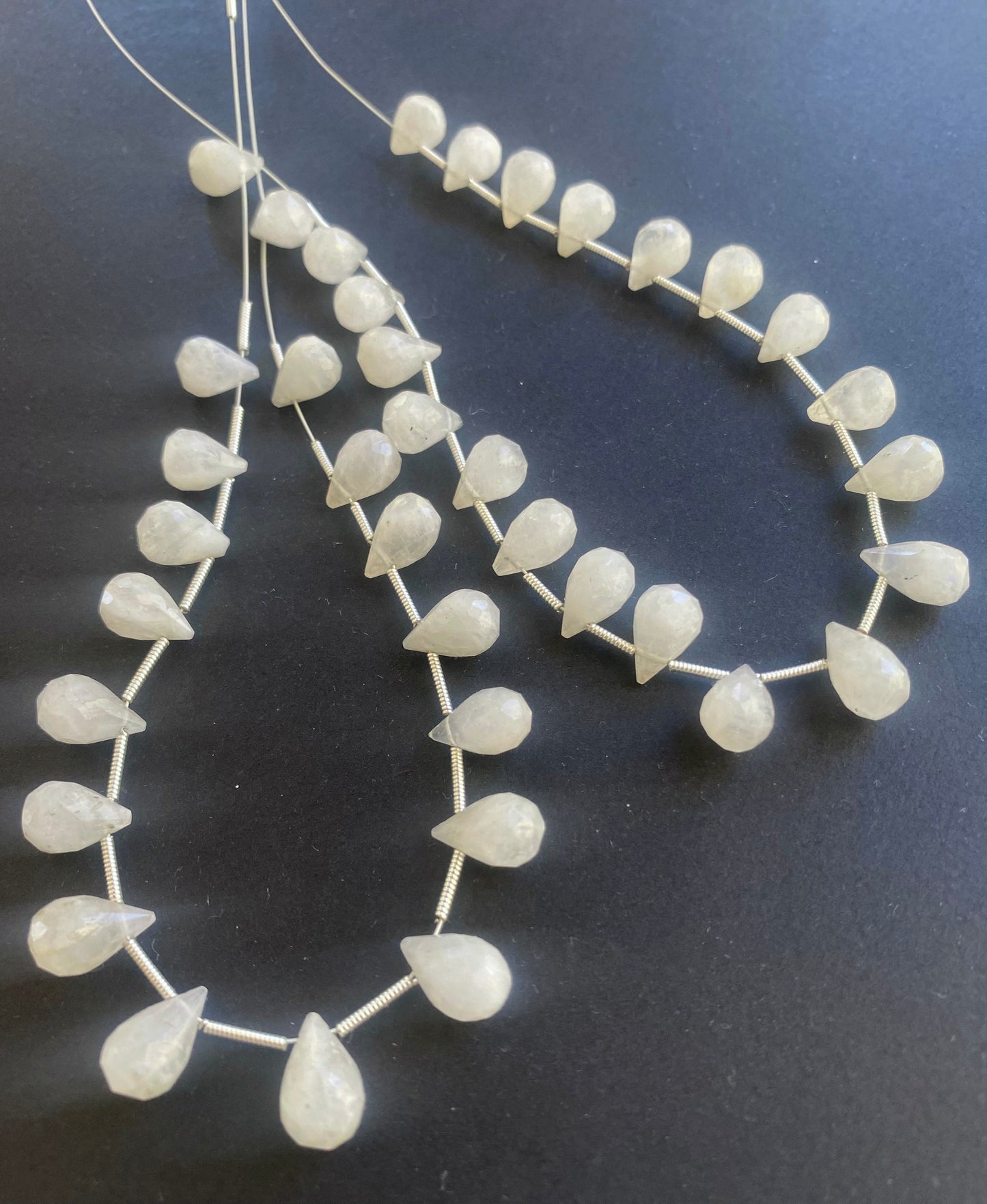 Strand of white teardrop shaped moonstone beads is displayed on a black surface, creating a striking contrast and visual appeal.