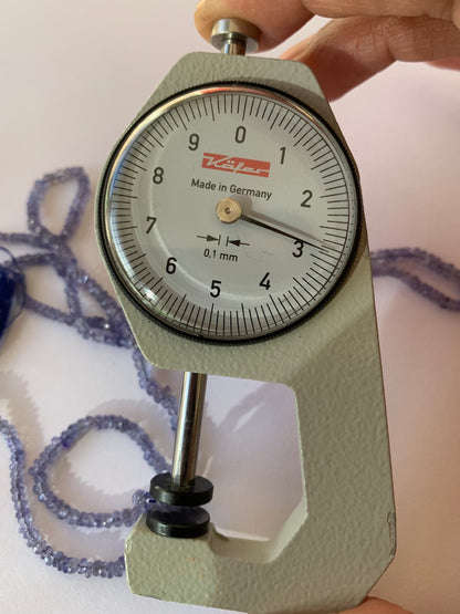A person measuring a Tanzanite faceted rondelle bead with a dial gauge measure, showcasing its size and detail.