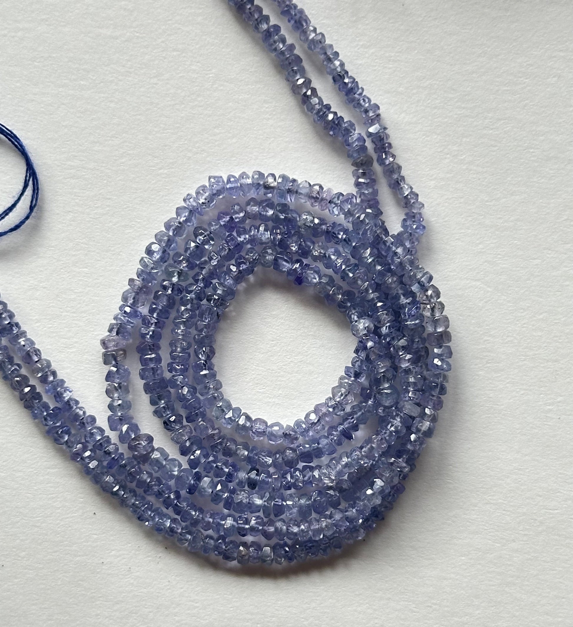 A close-up of a multiple strands of Tanzanite gemstone beads, faceted rondelles ranging from 2.4mm to 3mm, glistening in the light.