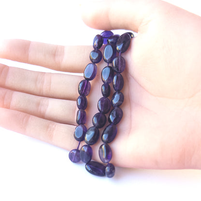 A person holds delicate strands of Amethyst beads, showcasing their smooth oval design.