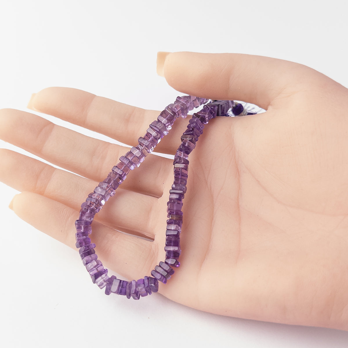 A person holds delicate strands of Amethyst beads, showcasing their smooth square heishi design.