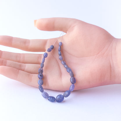 A person holds delicate strands of Blue Sapphire smooth oval beads, showcasing their polished design.