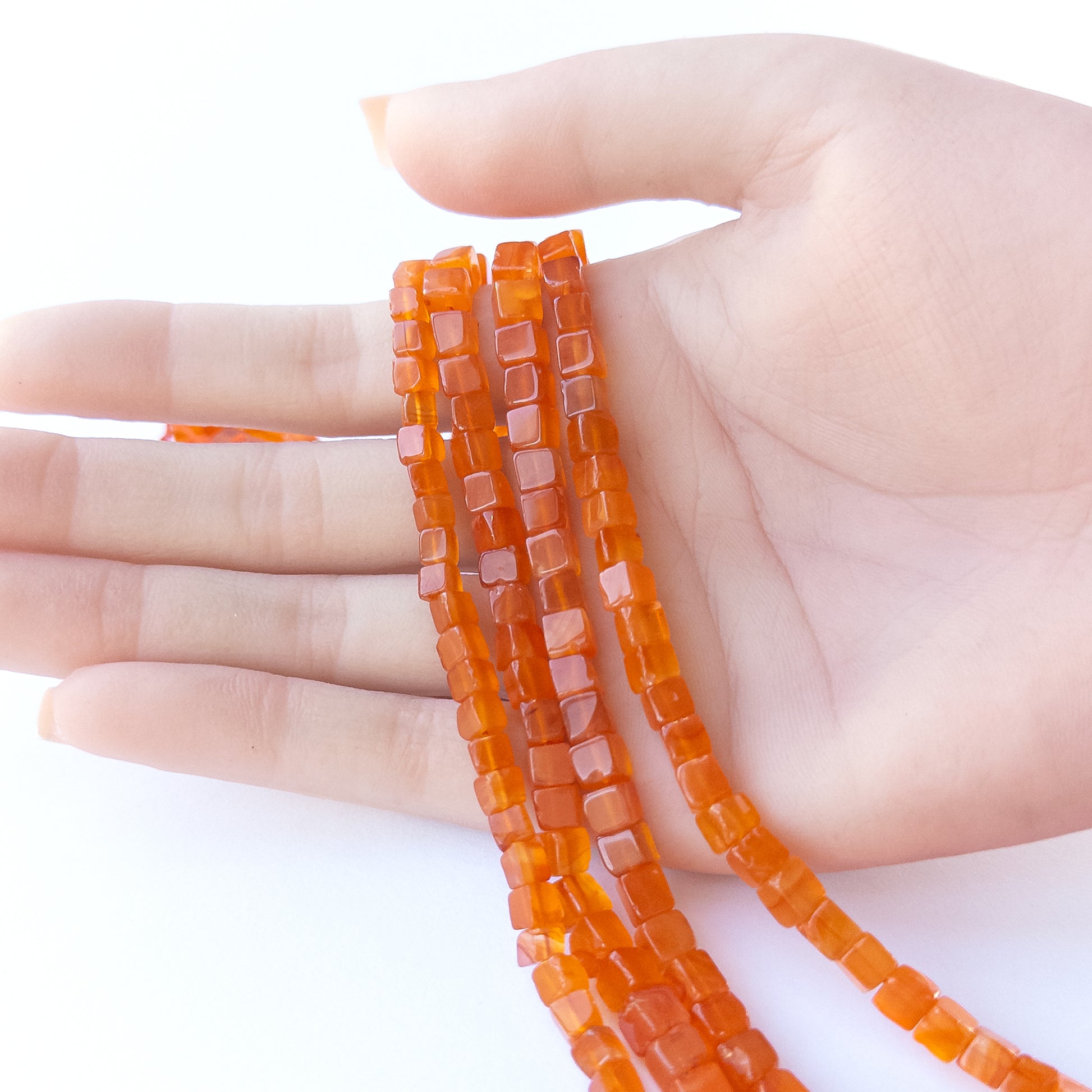 A person holds delicate strands of Carnelian beads, showcasing their smooth cube design.