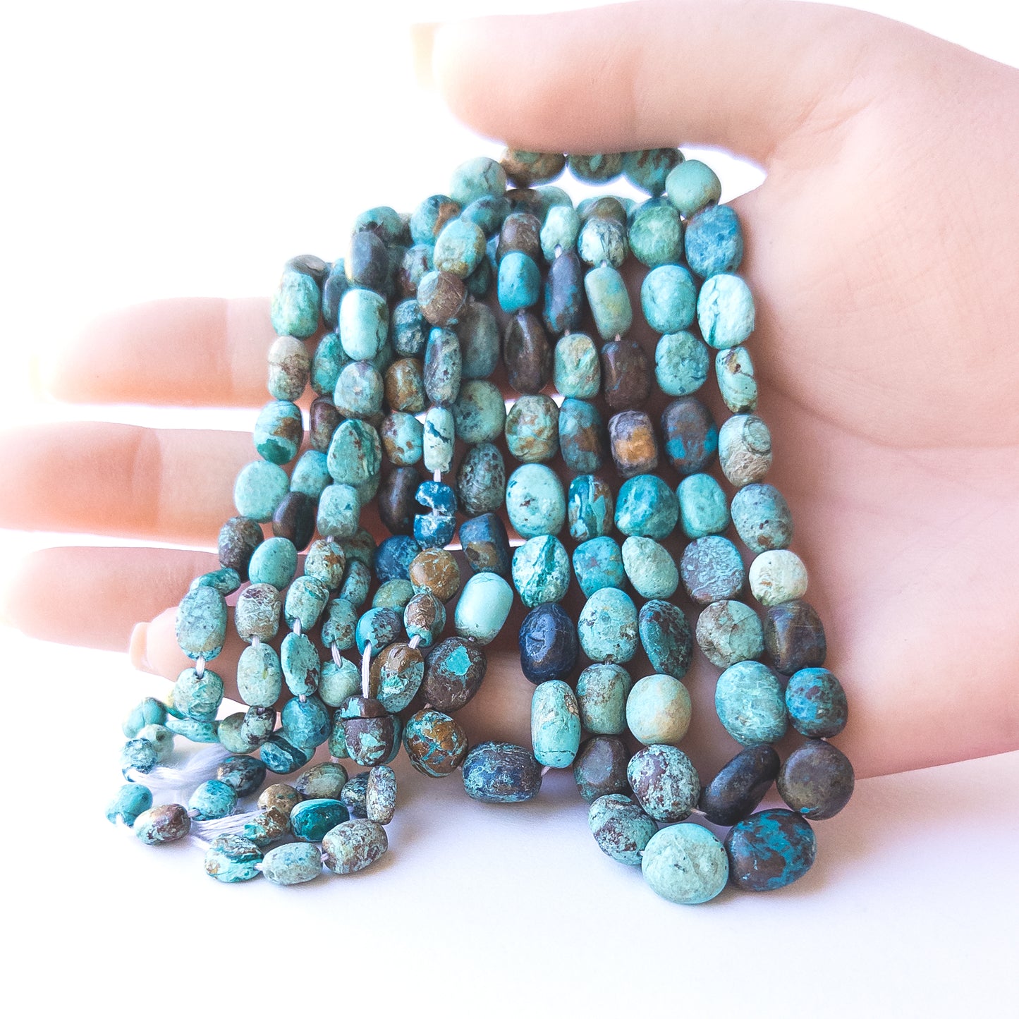 A person holds delicate strands of Chrysocolla beads, showcasing their smooth tumble design.  