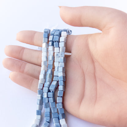 A person holds delicate strands of Denim Opal beads, showcasing their smooth cube design.