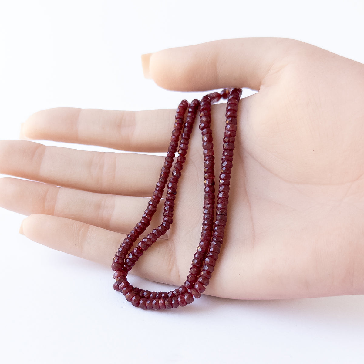 A person holds delicate strands of dyed Ruby beads, showcasing their faceted rondelle design.