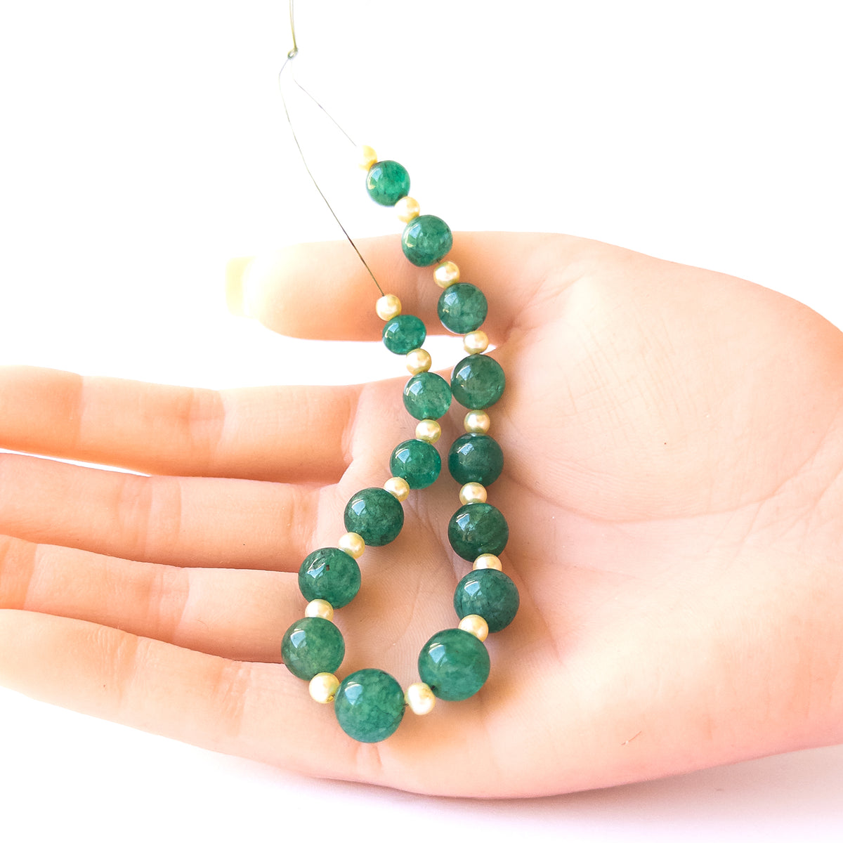 A person holds delicate pieces of Emerald and Shell Pearl beads, showcasing their smooth round design.