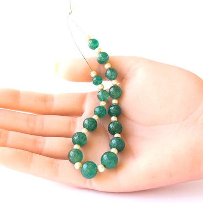 A person holds delicate pieces of Emerald and Shell Pearl beads, showcasing their smooth round design.
