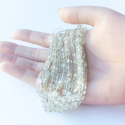A person holds delicate strands of Green Amethyst beads, showcasing their smooth cube design.