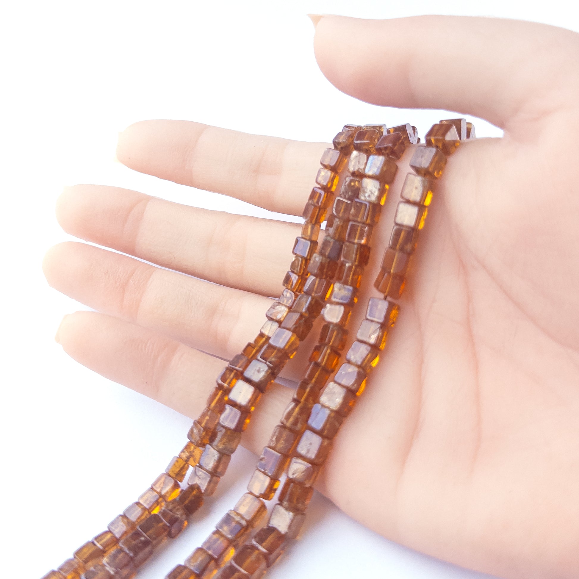 A person holds delicate strands of Hessonite beads, showcasing their smooth cube design.