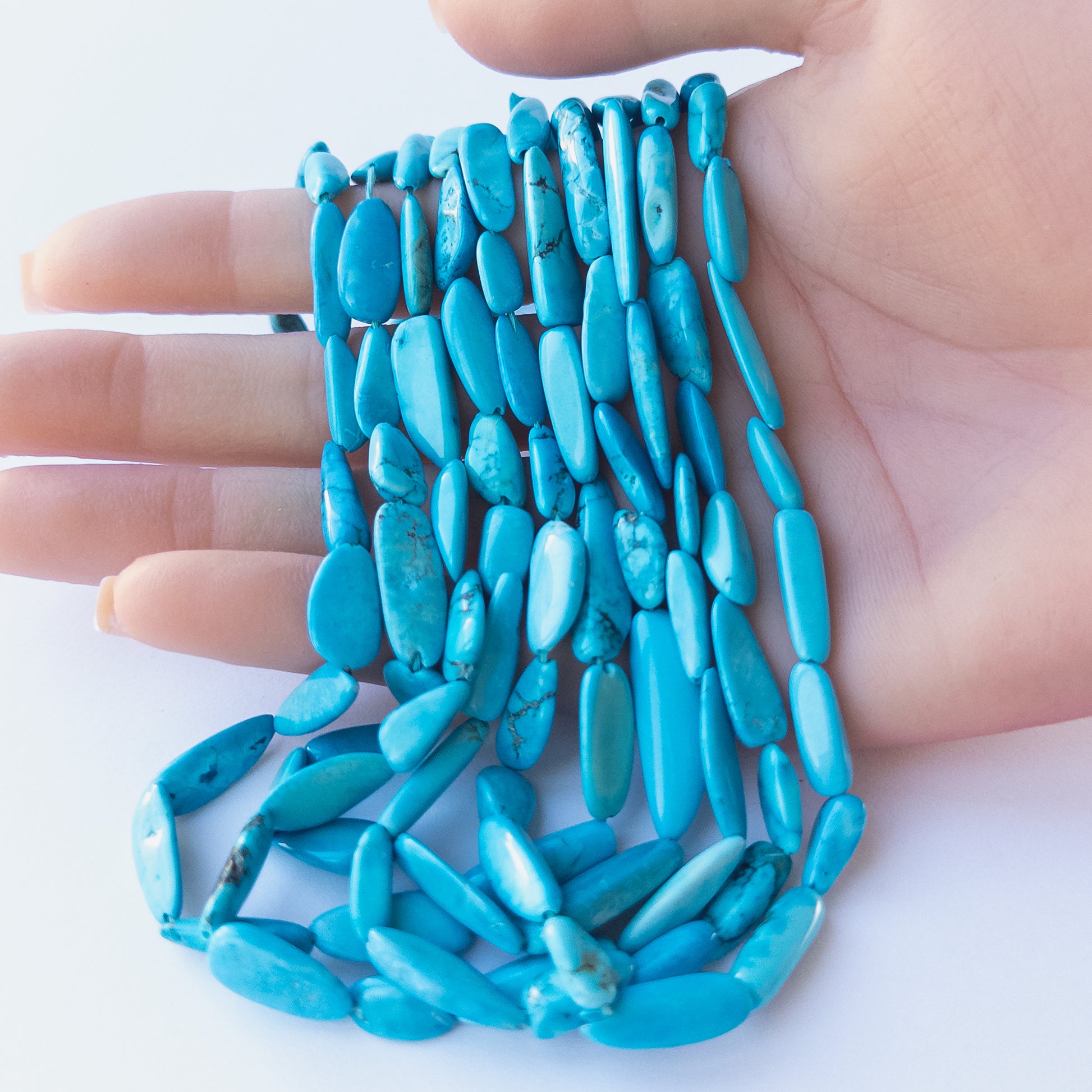 A person holds delicate strands of Howlite Turquoise beads, showcasing their smooth tumble design.