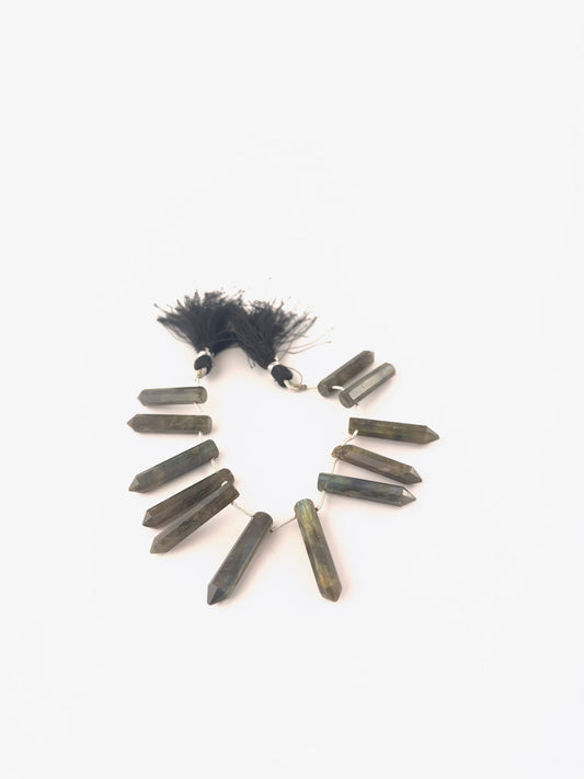 Labradorite faceted pencil briolette beads sized 6–7mm arranged as an 11‑piece set, showing grey‑blue body colour with flashes of labradorescence and sharp faceting.