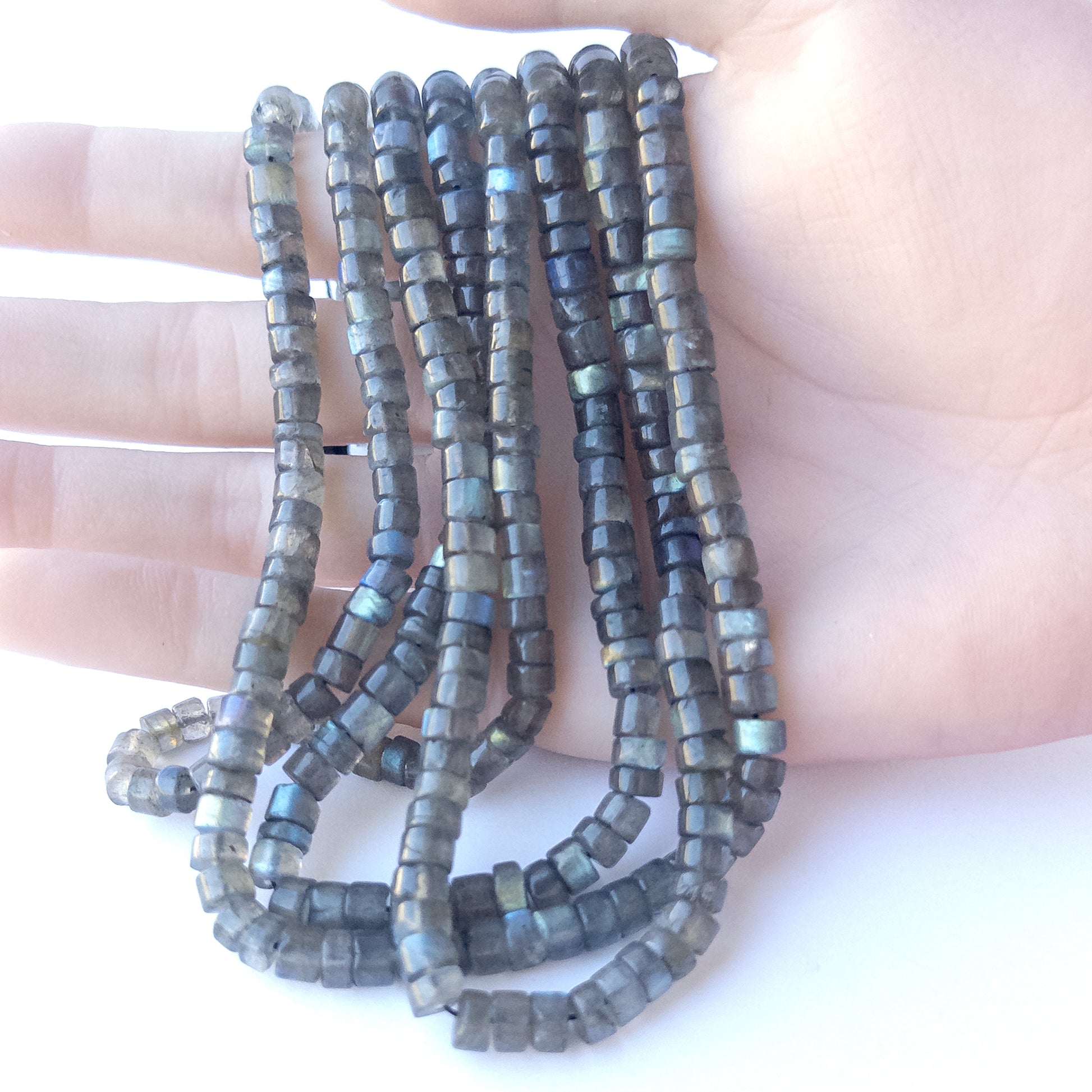 A person holds delicate strands of Labradorite beads, showcasing their smooth cylinder design.