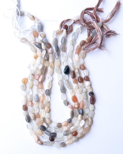 A person holds delicate strands of Multi Moonstone beads, showcasing their smooth tumble design.