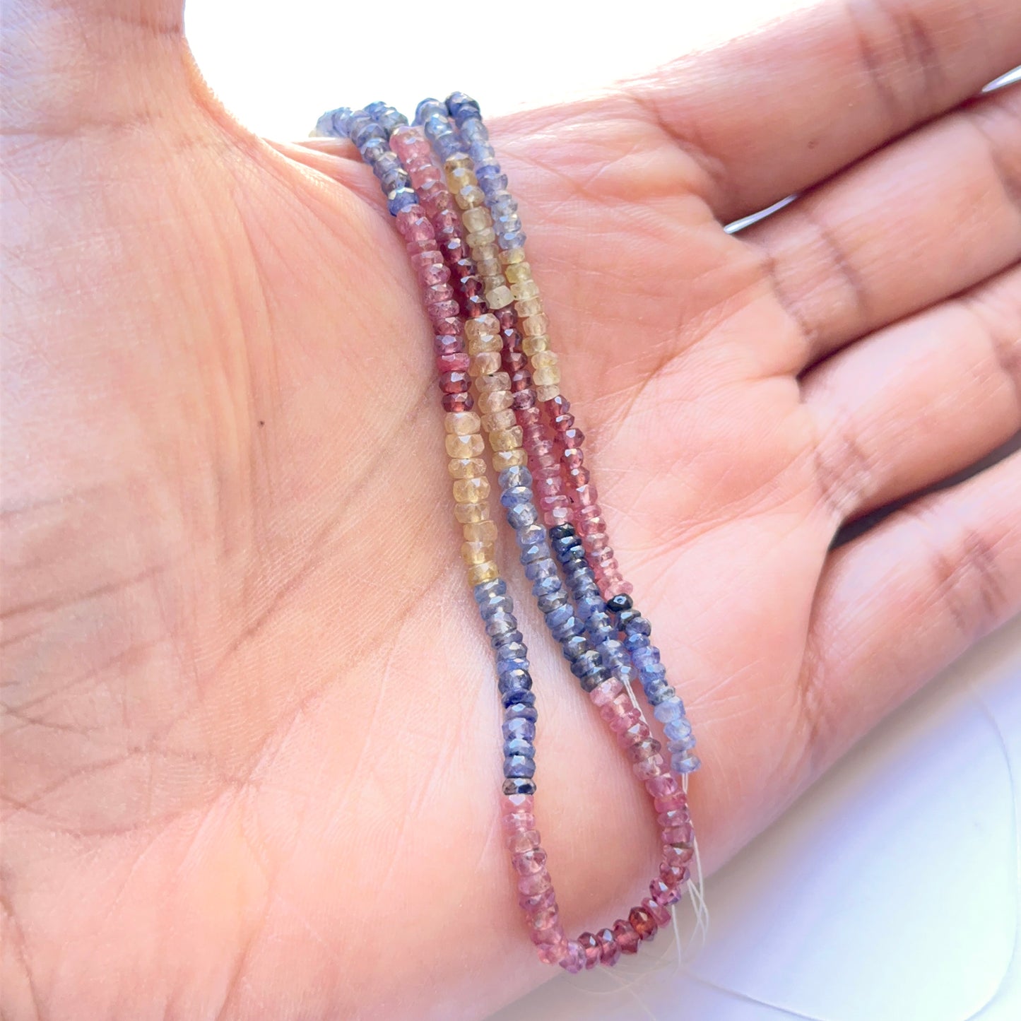 A person holds delicate strands of mixed precious faceted rondelle beads, showcasing ruby, yellow sapphire and blue sapphire.
