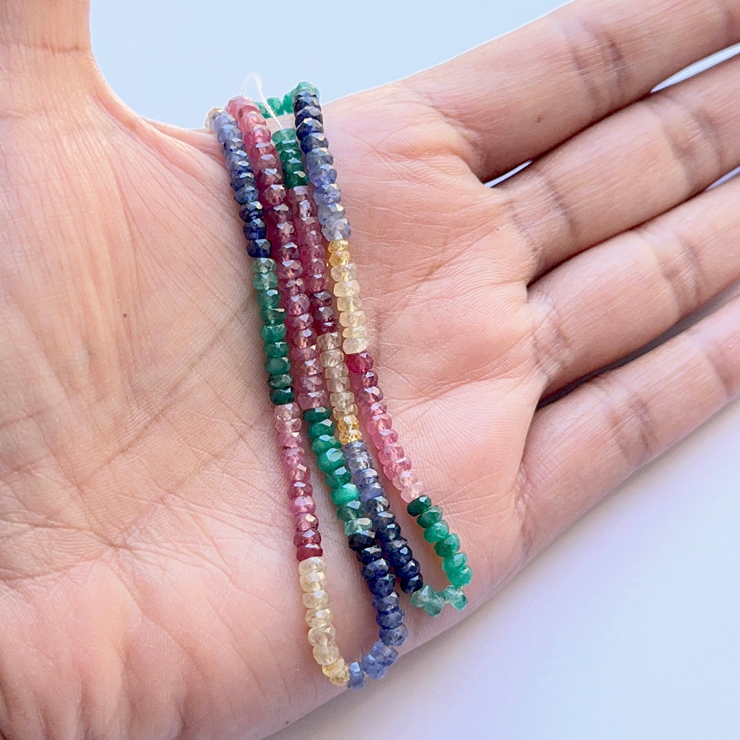 A person holds the mixed precious faceted rondelle strand, showcasing ruby, yellow sapphire, blue sapphire and emerald beads.

