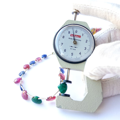 A dial gauge measures the Ruby, Blue Sapphire and Emerald faceted briolette 7–10mm gemstone.