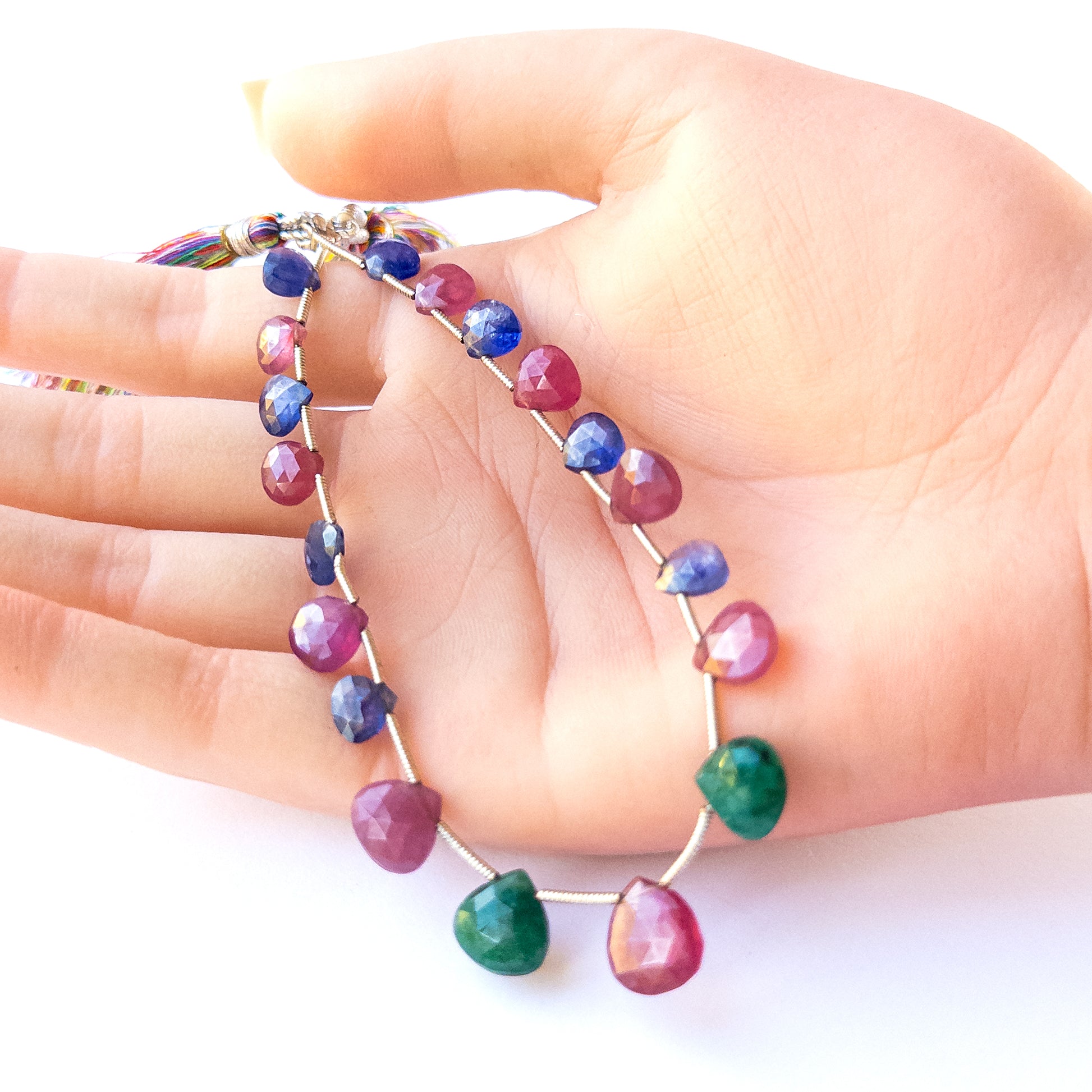 A person holds delicate strands of Ruby, Blue Sapphire and Emerald briolette beads, showcasing their design.