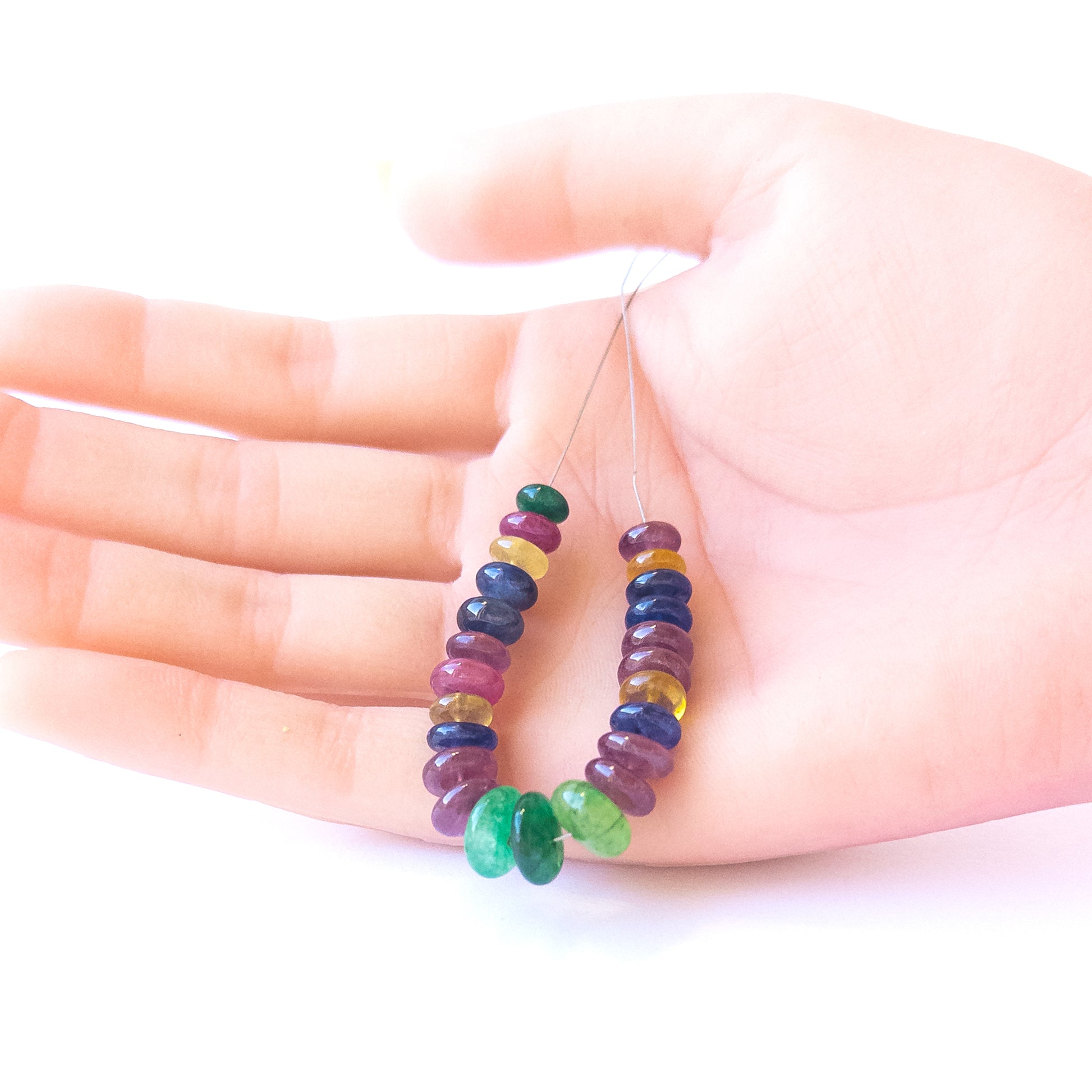 A person holds delicate strands of Ruby, Emerald and Sapphire smooth rondelle beads, showcasing their polished design.  
