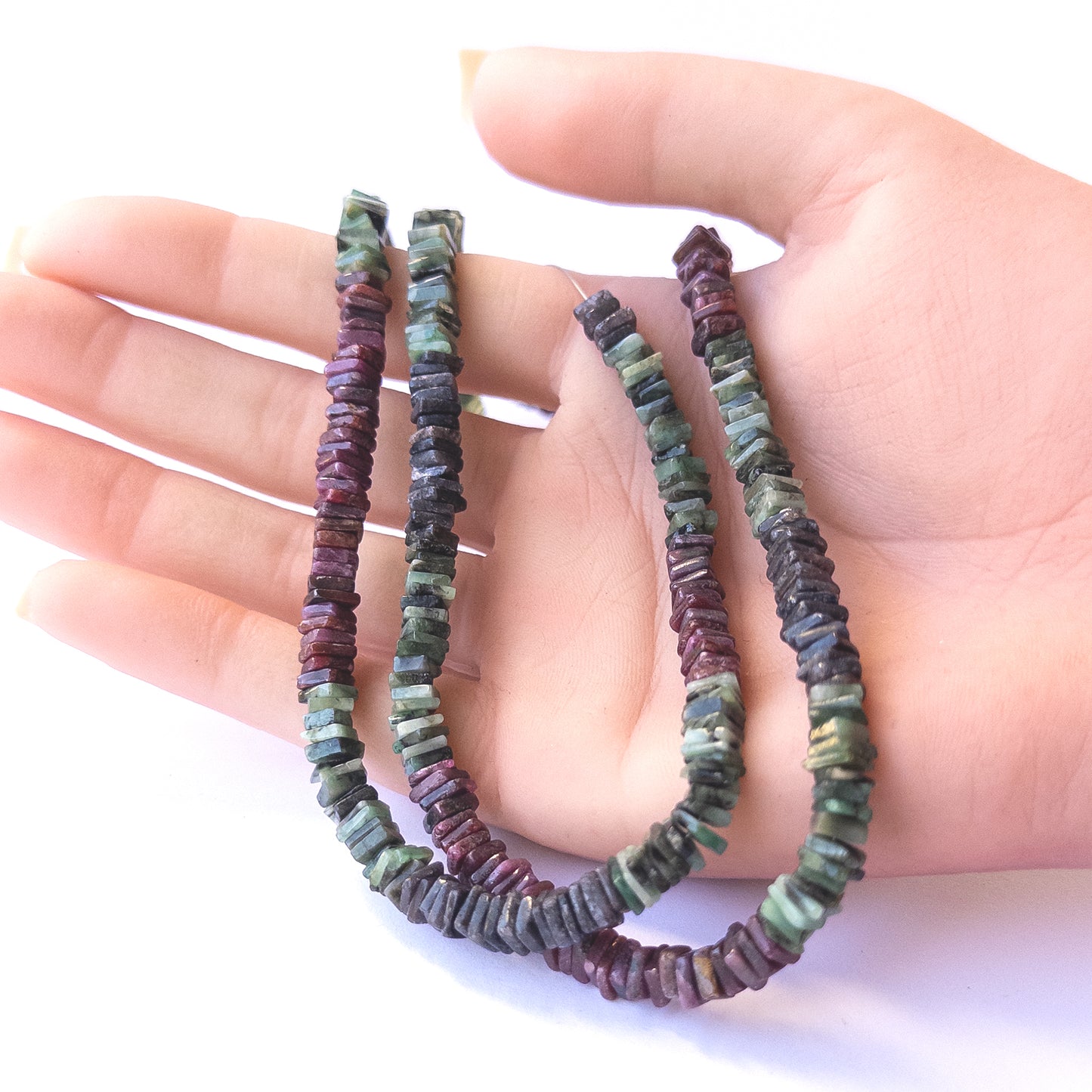 A person holds delicate strands of Ruby, Sapphire and Emerald beads, showcasing their smooth square heishi design.