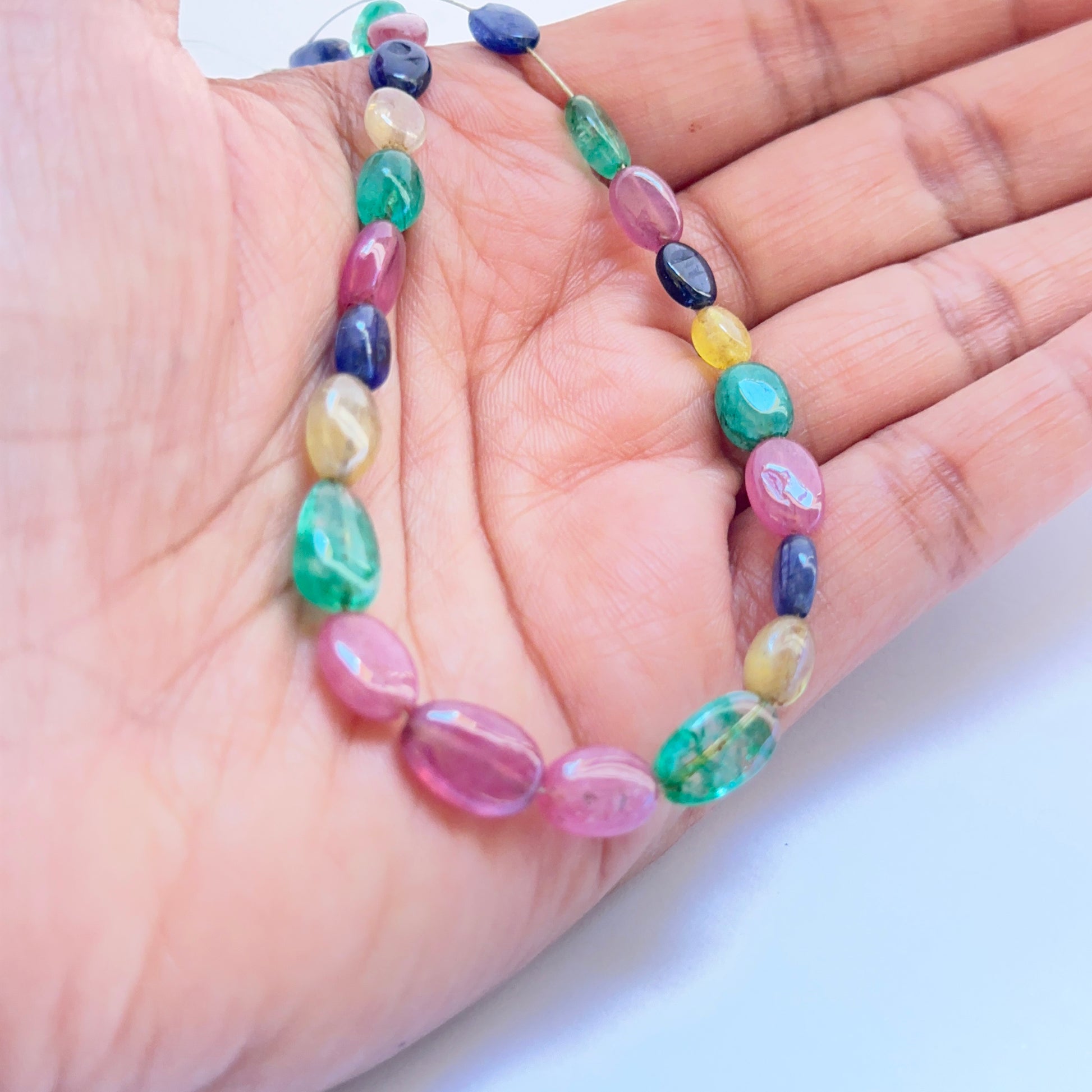 A person holds the multi stone smooth oval bead set, showcasing ruby, blue sapphire and emerald tones.