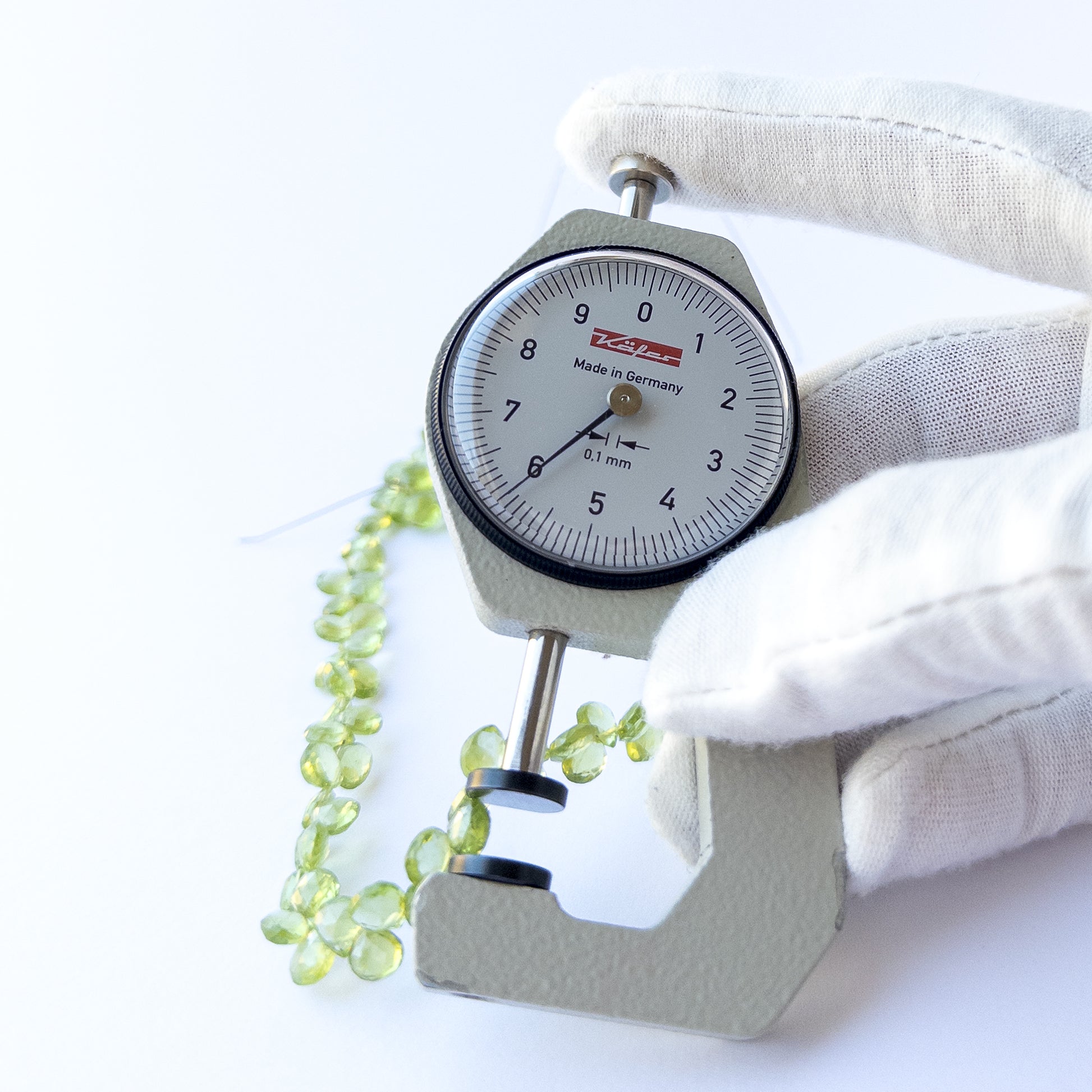 A dial gauge measures the Peridot faceted briolette 5.5–6mm gemstone.
