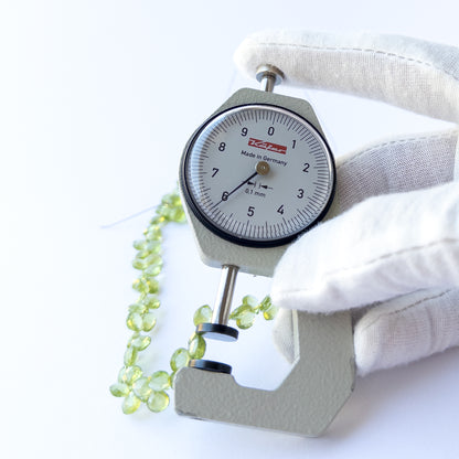 A dial gauge measures the Peridot faceted briolette 5.5–6mm gemstone.