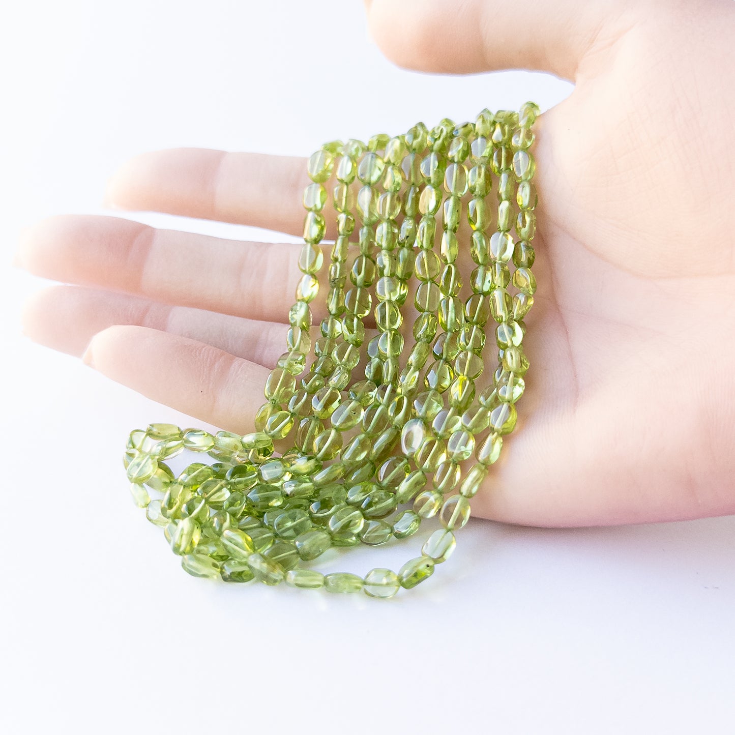 A person holds delicate strands of Peridot beads, showcasing their smooth oval design.
