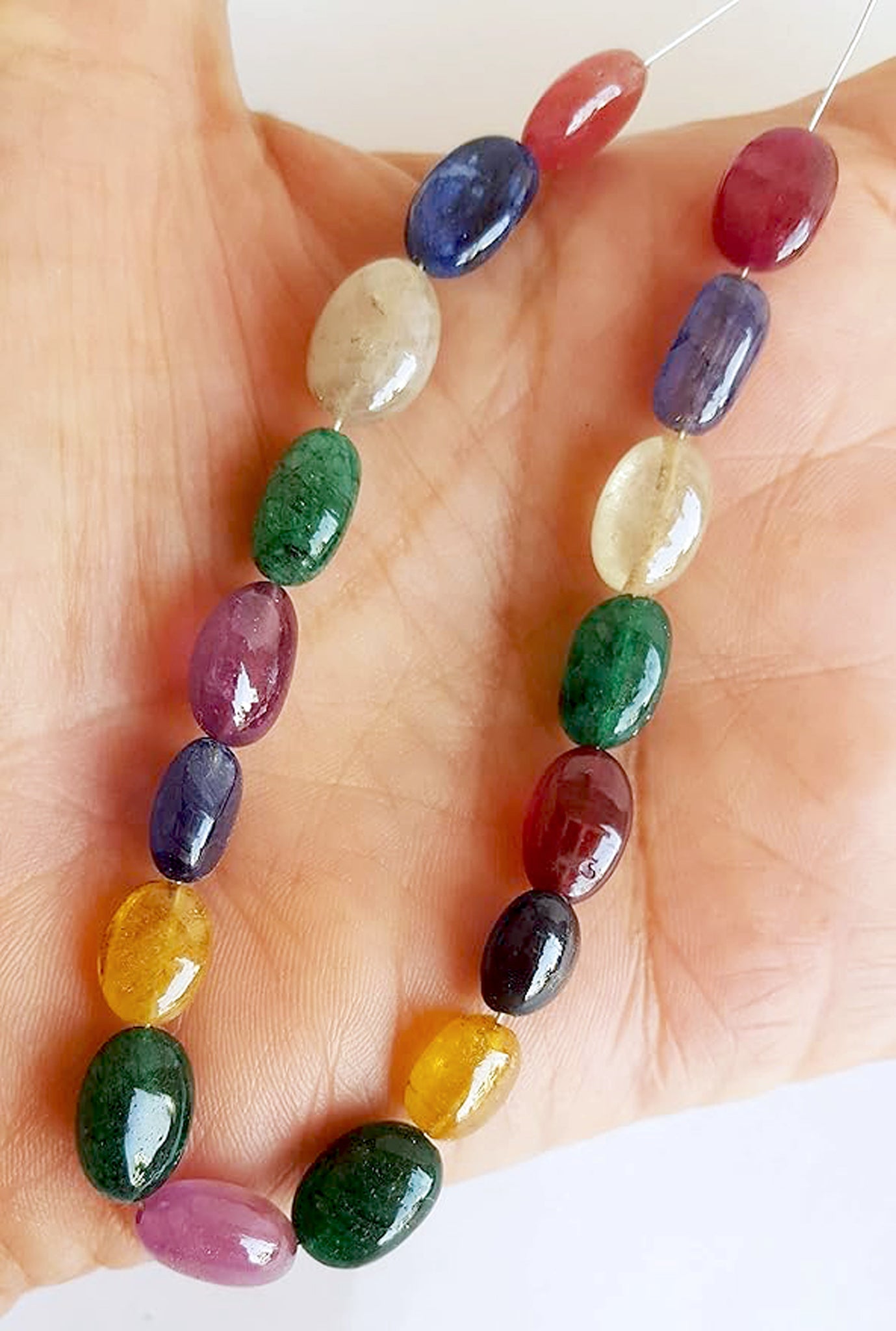  A person holds a vibrant bunch of multi-coloured smooth oval beads, including ruby, blue sapphire, yellow sapphire, and emerald.