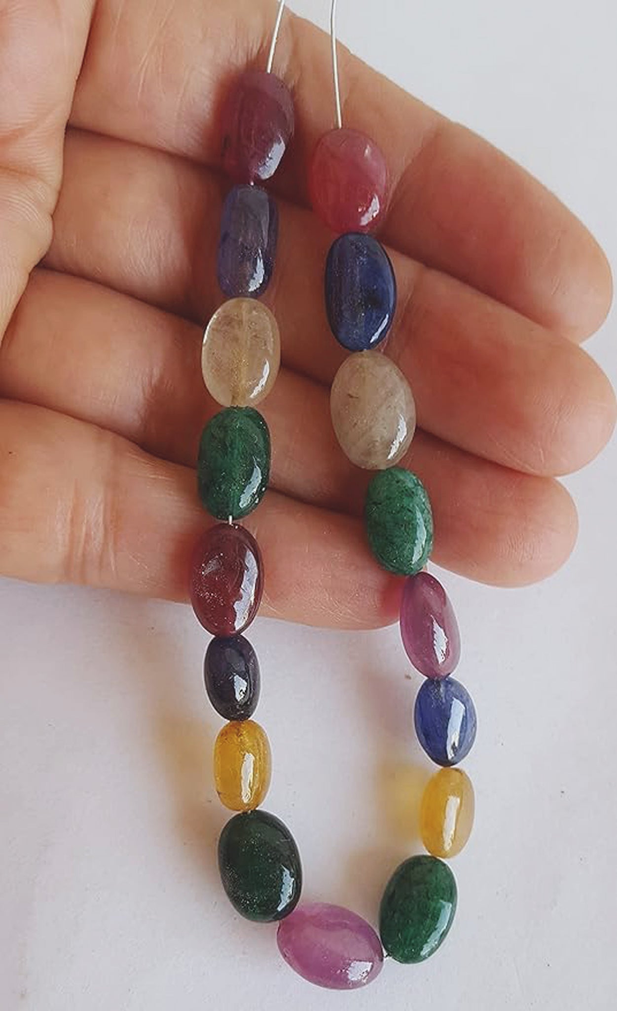  A person holds a vibrant bunch of multi-coloured smooth oval beads, including ruby, blue sapphire, yellow sapphire, and emerald.