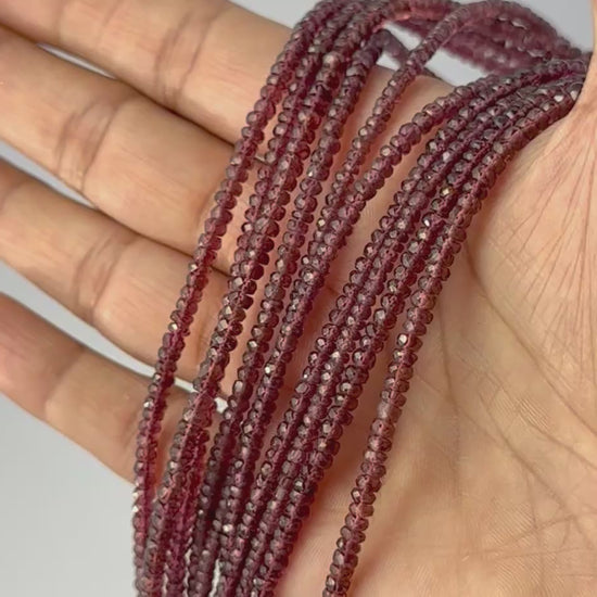 A video person holds delicate strands of Burma Garnet beads, showcasing their faceted rondelle design and sparkle.