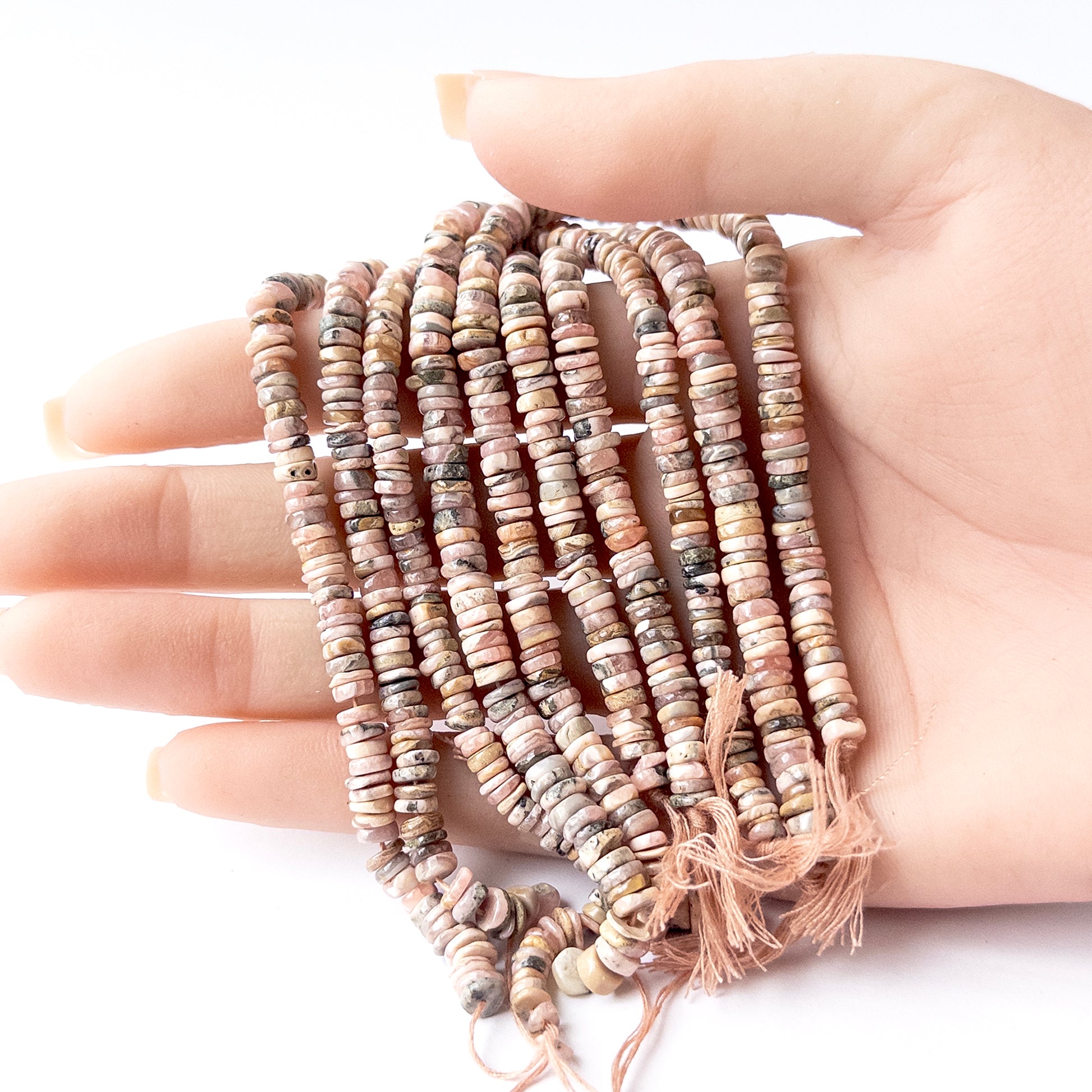 A person holds delicate strands of Rhodonite beads, showcasing their smooth wheel heishi design.