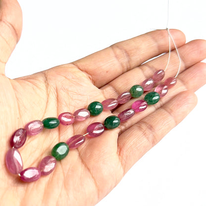 A person holds delicate strands of ruby and emerald oval beads, showcasing their smooth design.