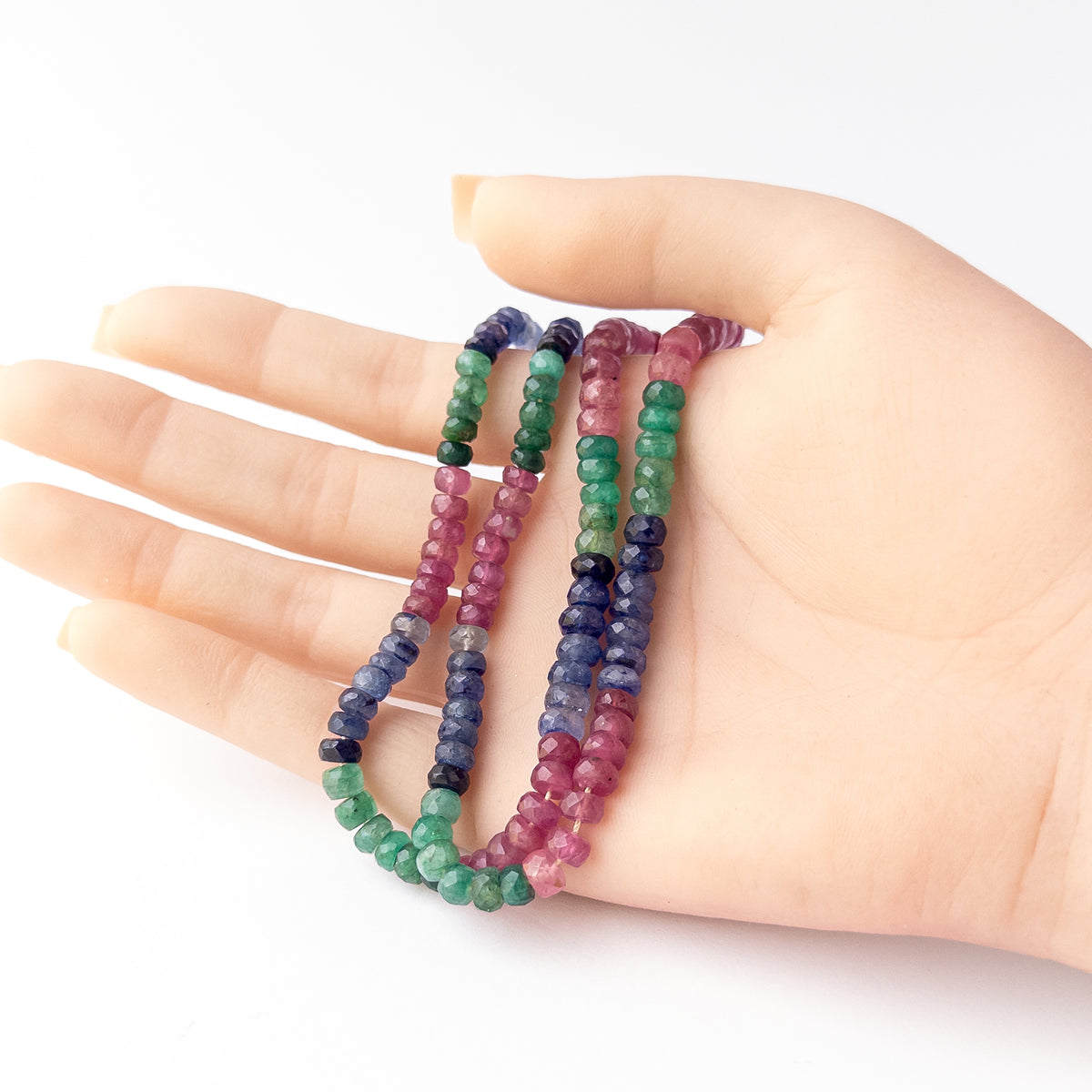 A person holds delicate strands of Multi Precious Ruby Blue Sapphire and Emerald beads, showcasing their faceted rondelle design