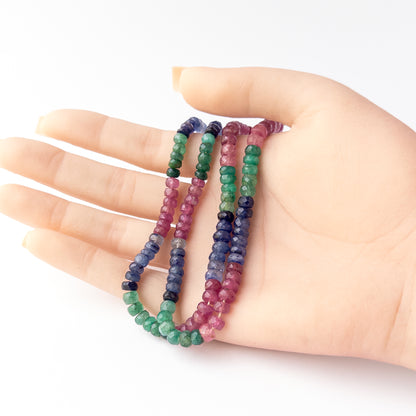 A person holds delicate strands of Multi Precious Ruby Blue Sapphire and Emerald beads, showcasing their faceted rondelle design
