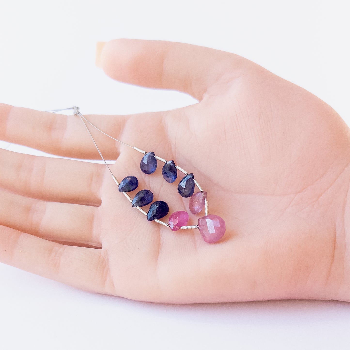 A person holds delicate strands of Ruby and Blue Sapphire beads, showcasing their faceted briolette design.  
