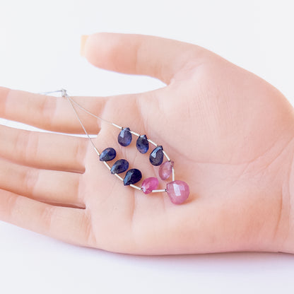 A person holds delicate strands of Ruby and Blue Sapphire beads, showcasing their faceted briolette design.  
