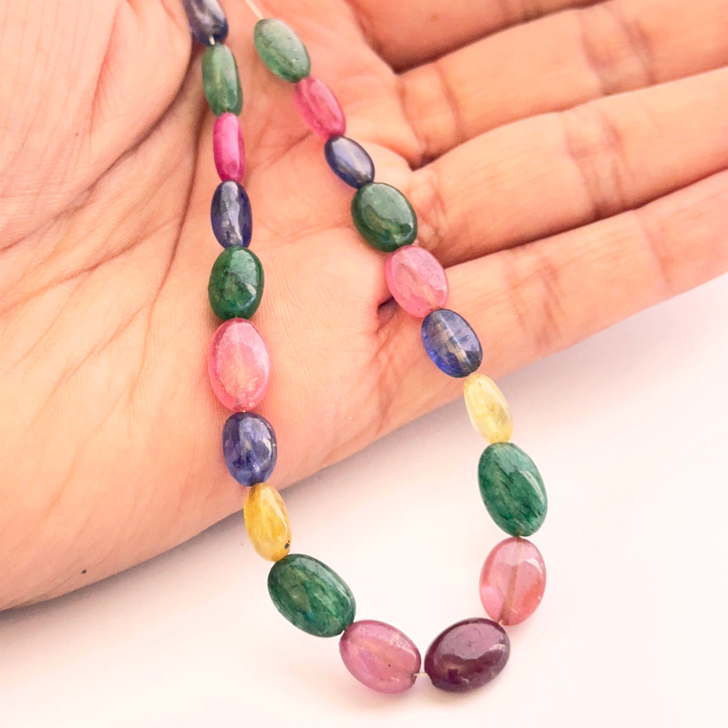 A person holds the multi stone smooth oval bead set, showcasing ruby, blue sapphire, yellow sapphire and emerald.
