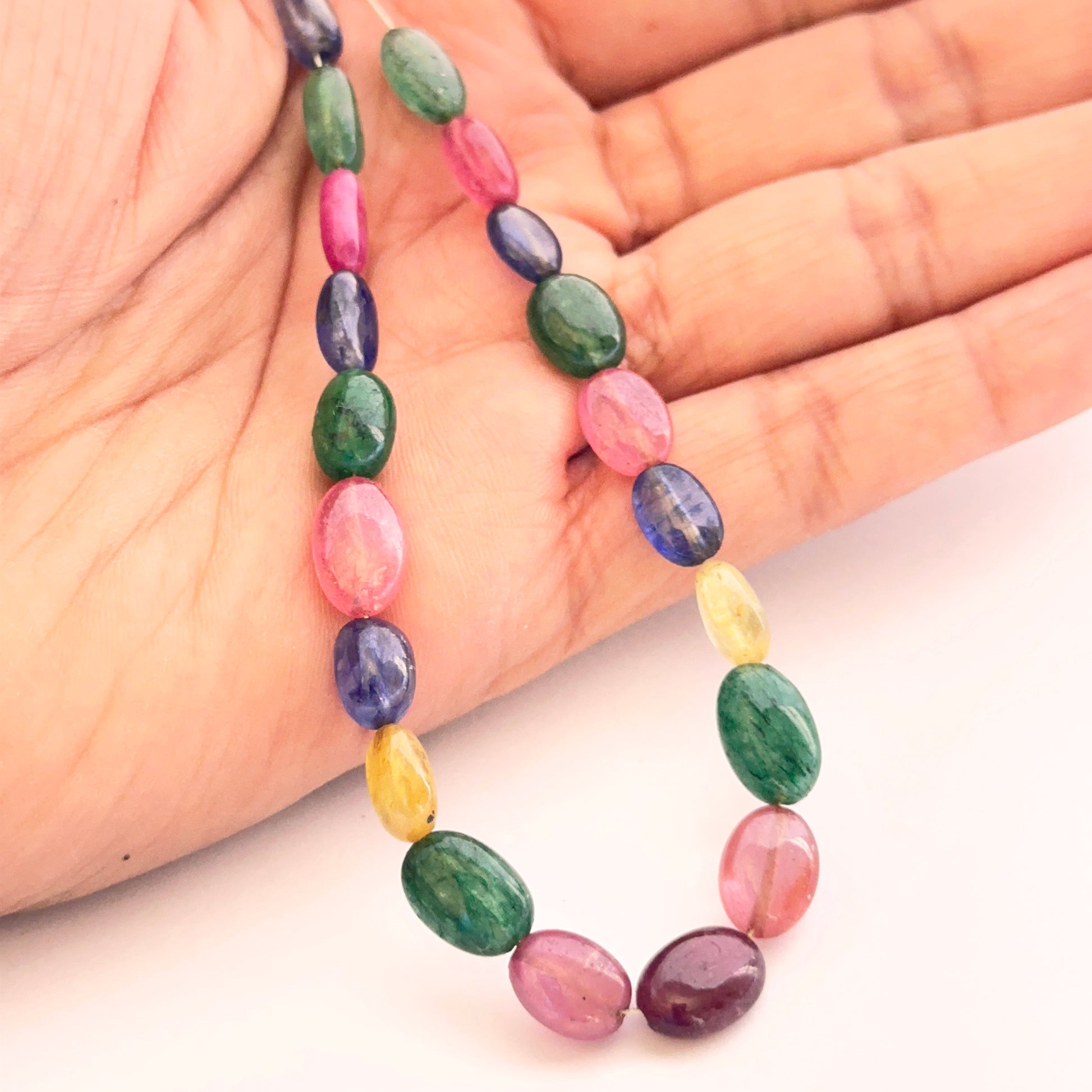 A person holds the multi stone smooth oval bead set, showcasing ruby, blue sapphire, yellow sapphire and emerald.
