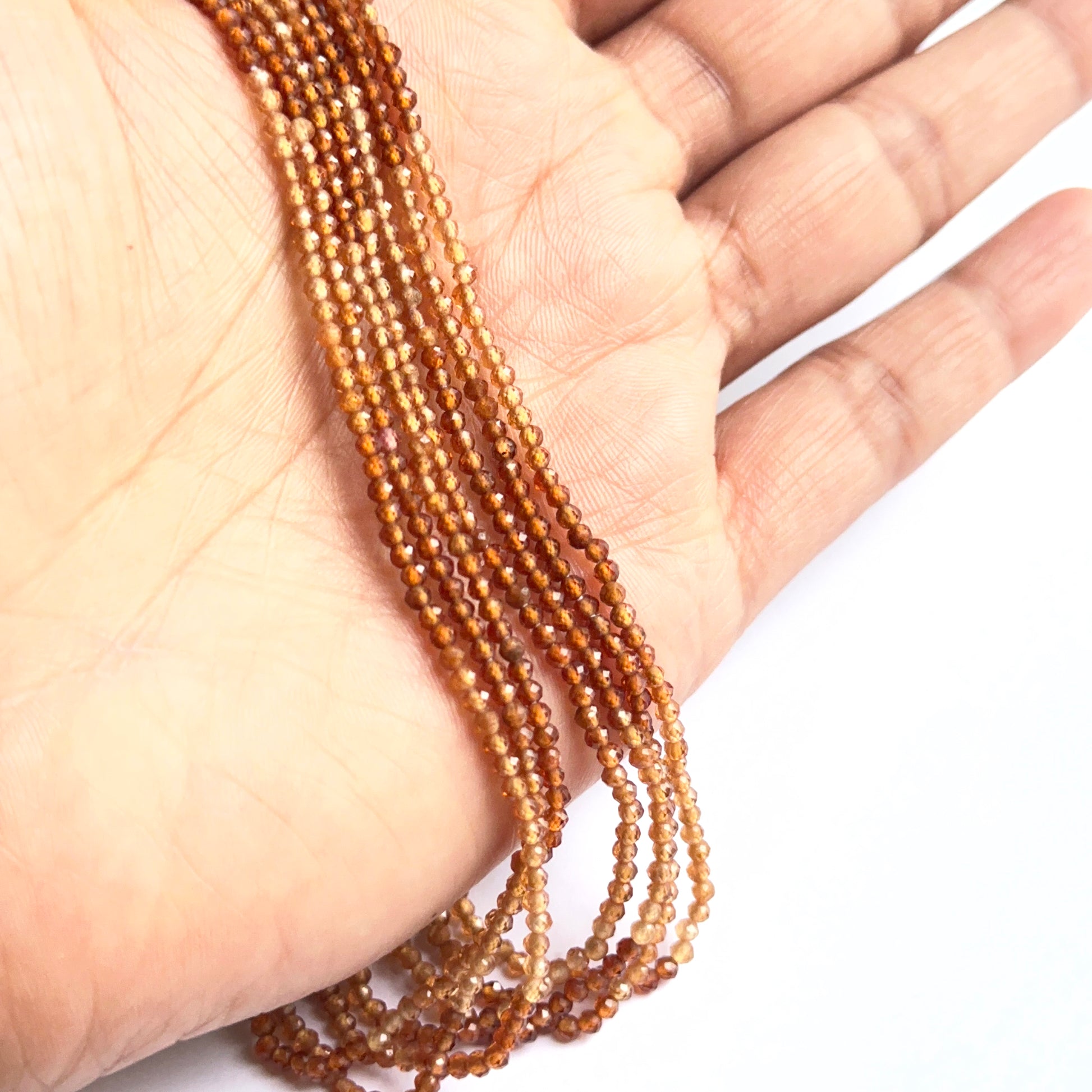 A person holds delicate strands of shaded hessonite faceted beads, showcasing their smooth design.