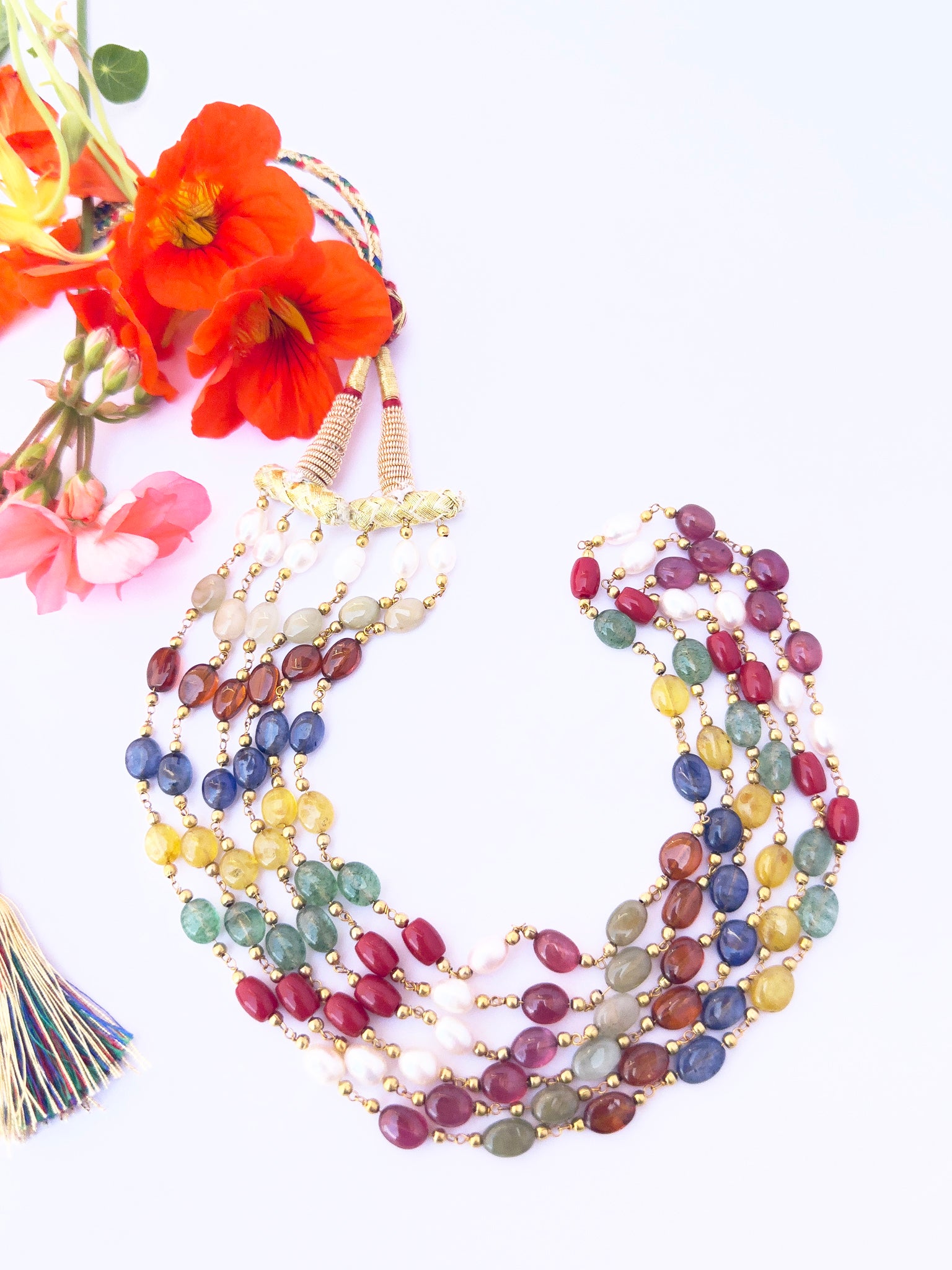 Navratan stone beads necklace, set next to orange and pink flowers, showcasing smooth oval design, 51–54cm adjustable length.