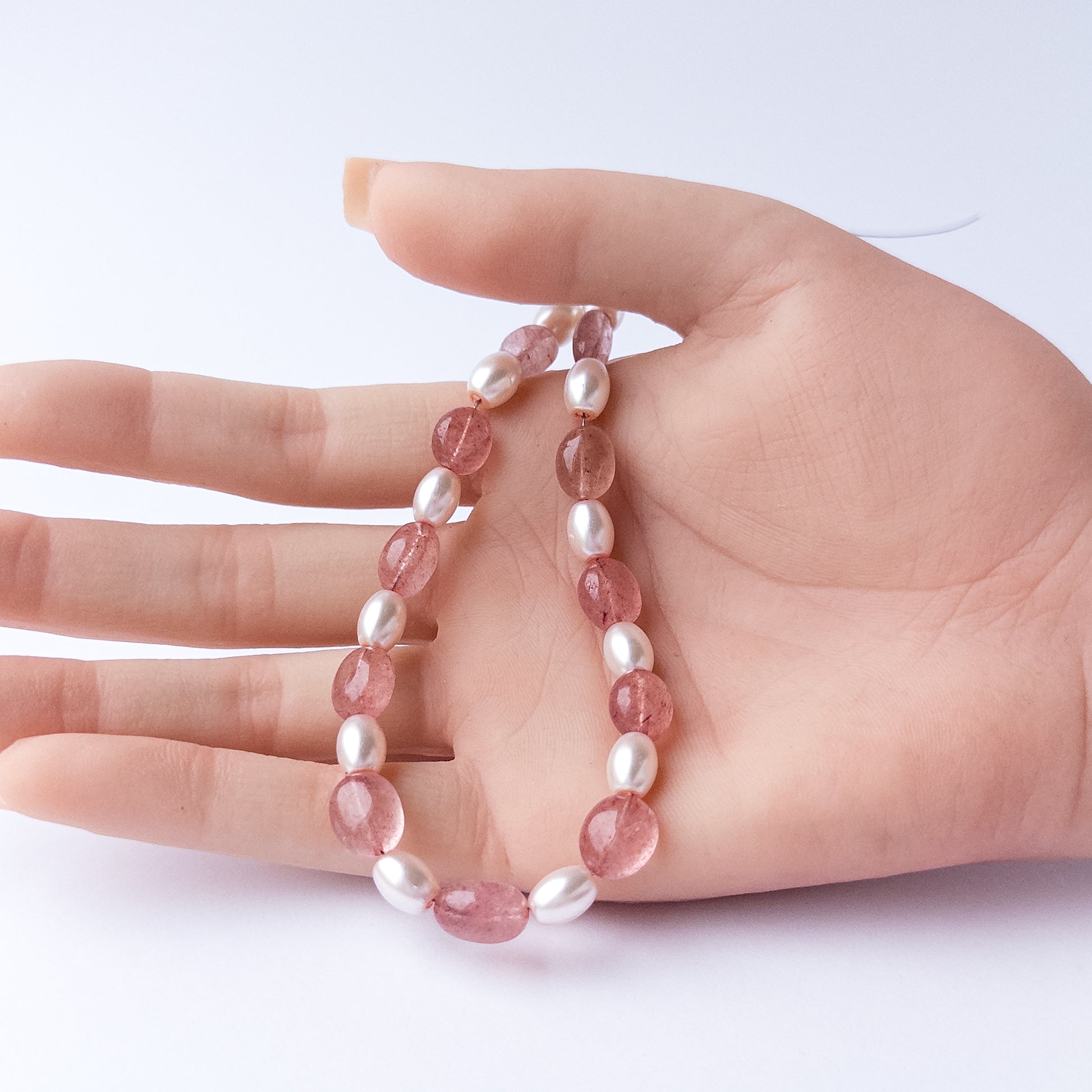 A person holds delicate strands of Strawberry Quartz and Shell Pearl beads, showcasing their smooth oval design.