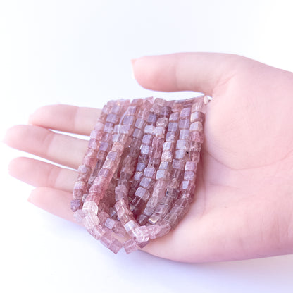 A person holds delicate strands of Strawberry Quartz beads, showcasing their smooth cube design.