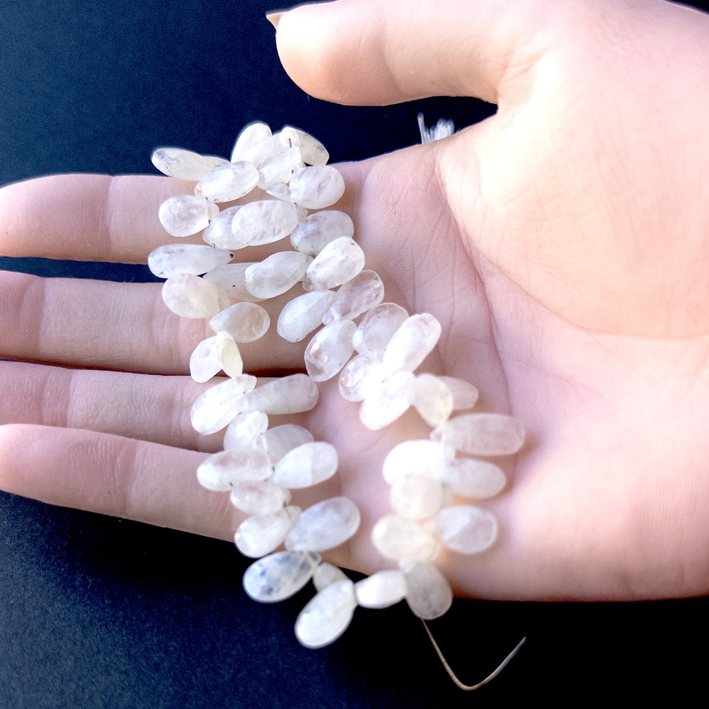 A person holds delicate strands of White Rainbow Moonstone faceted briolette pear beads, showcasing their sparkling design.