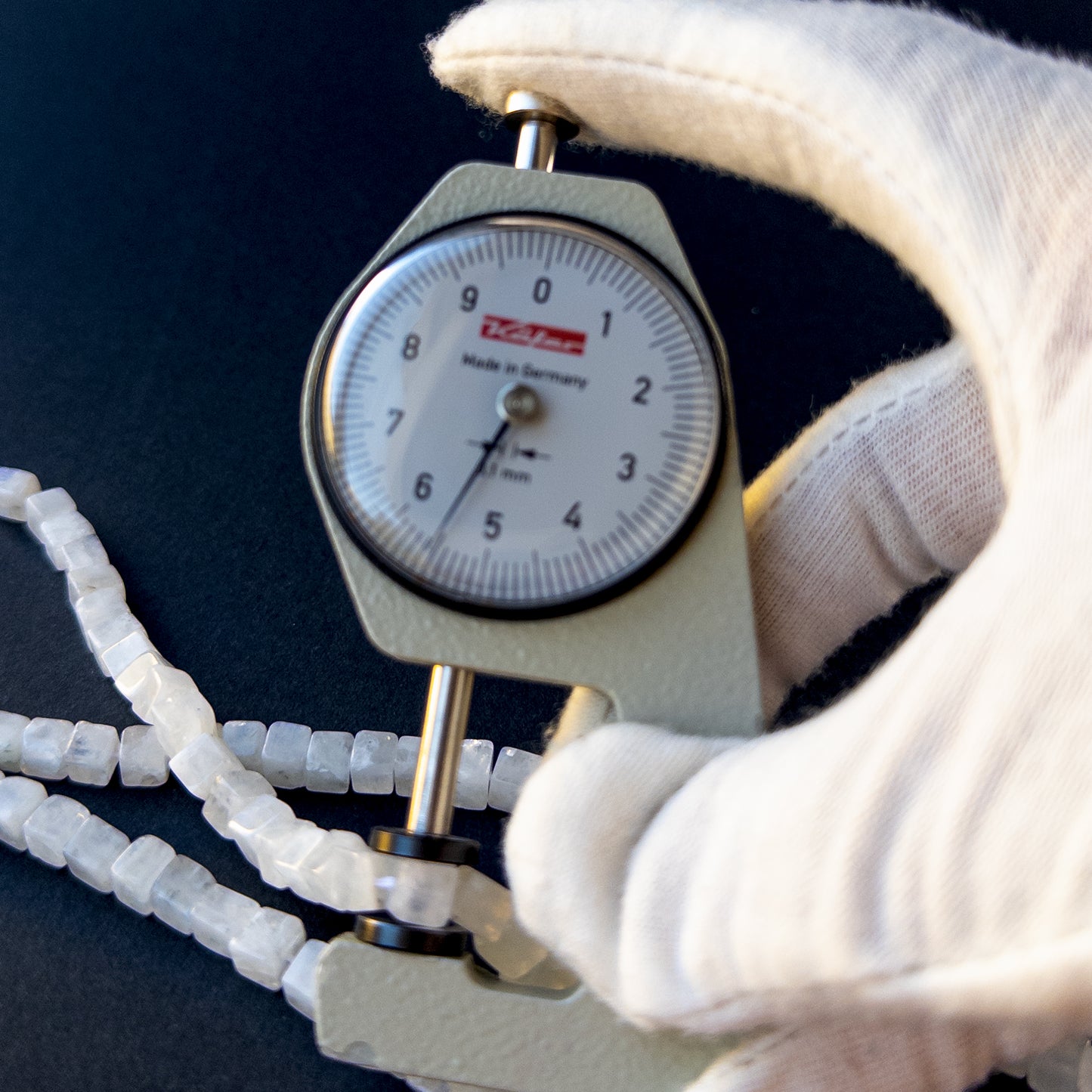 A dial gauge measures the White Rainbow Moonstone smooth cube 4.7–5.5mm gemstone.