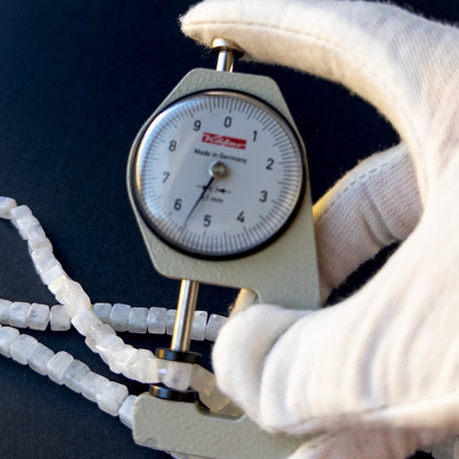 A dial gauge measures the White Rainbow Moonstone smooth cube 4.7–5.5mm gemstone.